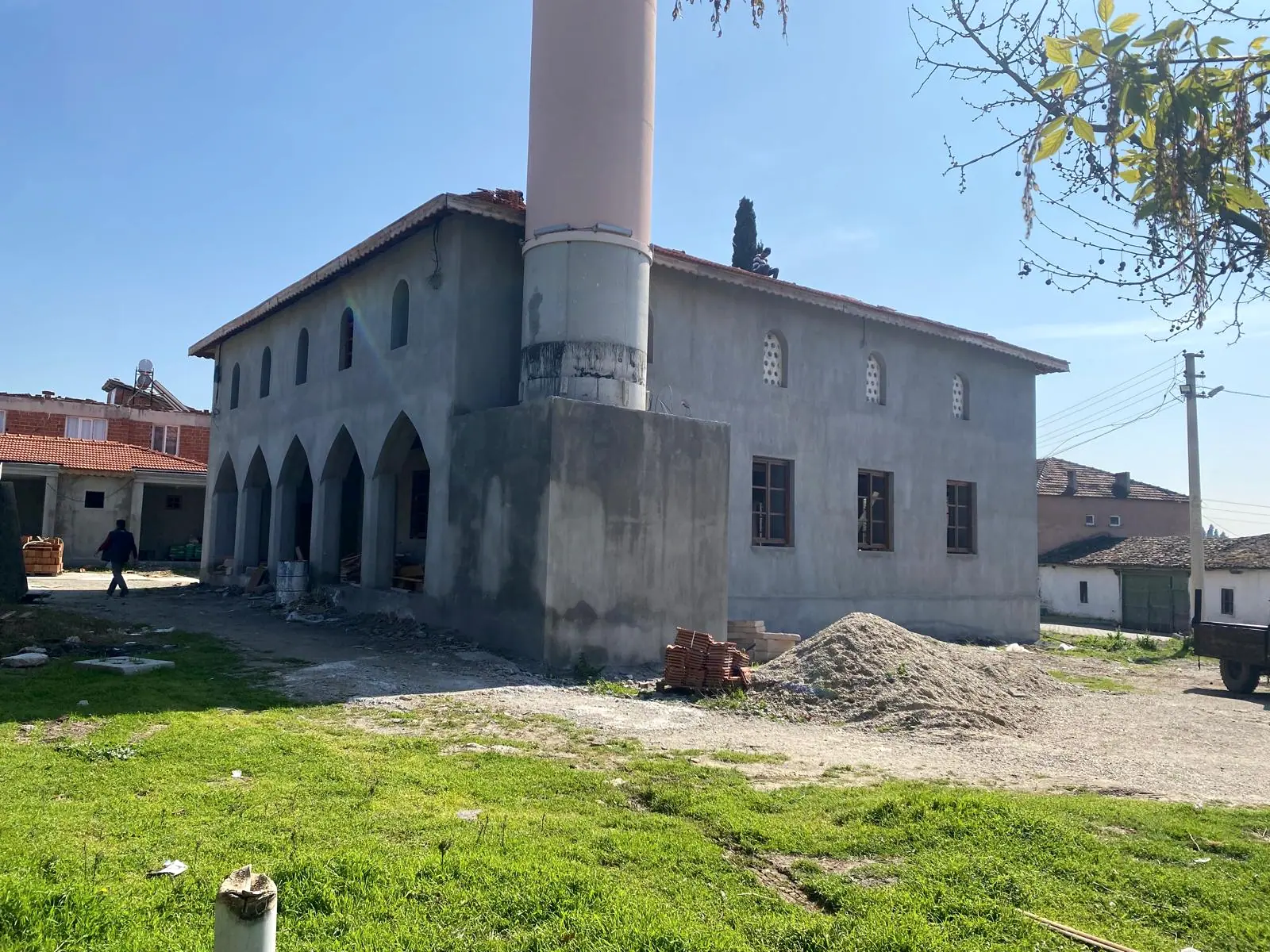 Cami arka köşesi: sıvalı duvarlar, ahşap doğrama pencereler, minare kaidesi, kum yığını ve paketli kiremitler