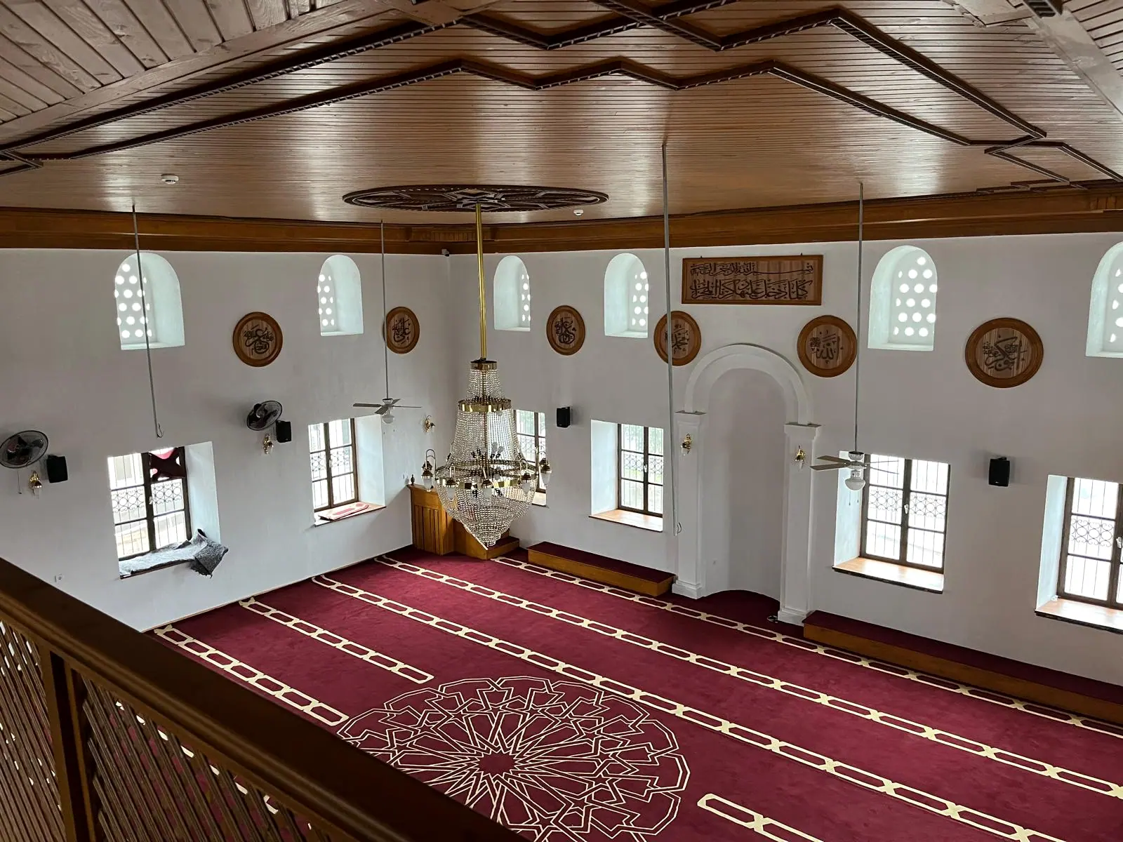 Mahfilden ana ibadet alanı: oyma göbekli ahşap tavan, kristal avize, hat levhaları ve saf çizgili halı