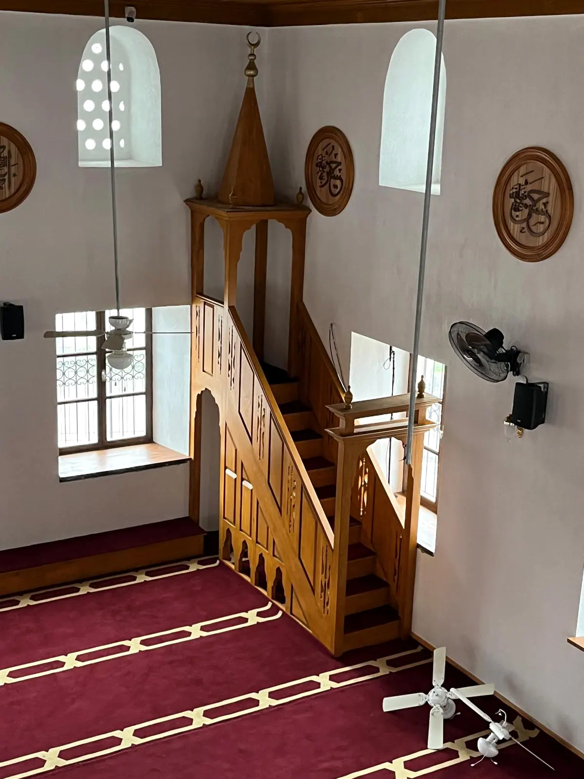 Oyma ahşap minber, konik külahı ve alemi, bordo halı basamak kaplaması ve duvardaki ahşap hat madalyonları