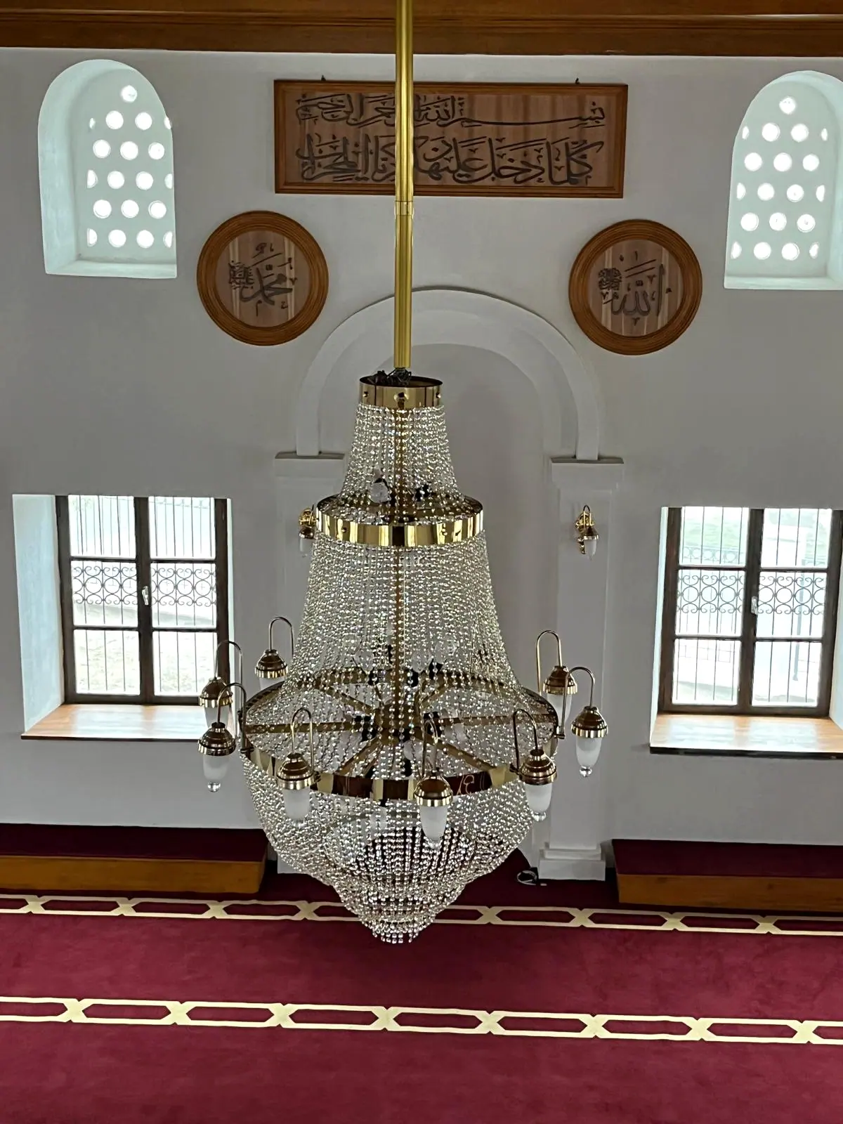 Kemerli mihrap nişi önünde asılı kristal ve pirinç avize, hat levhası ve ferforje korkuluklu pencereler