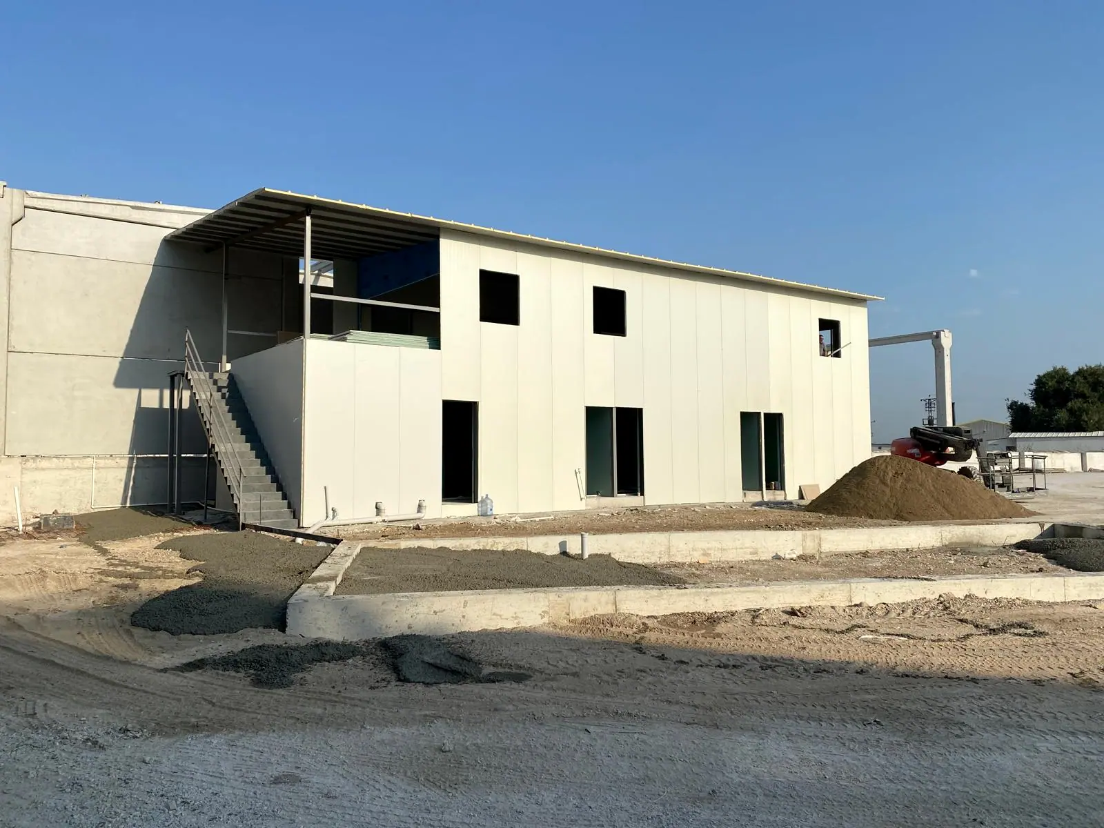 Beyaz sandviç panel cepheli iki katlı idari blok, dış çelik merdiven ve önünde taze beton dökülmüş saha