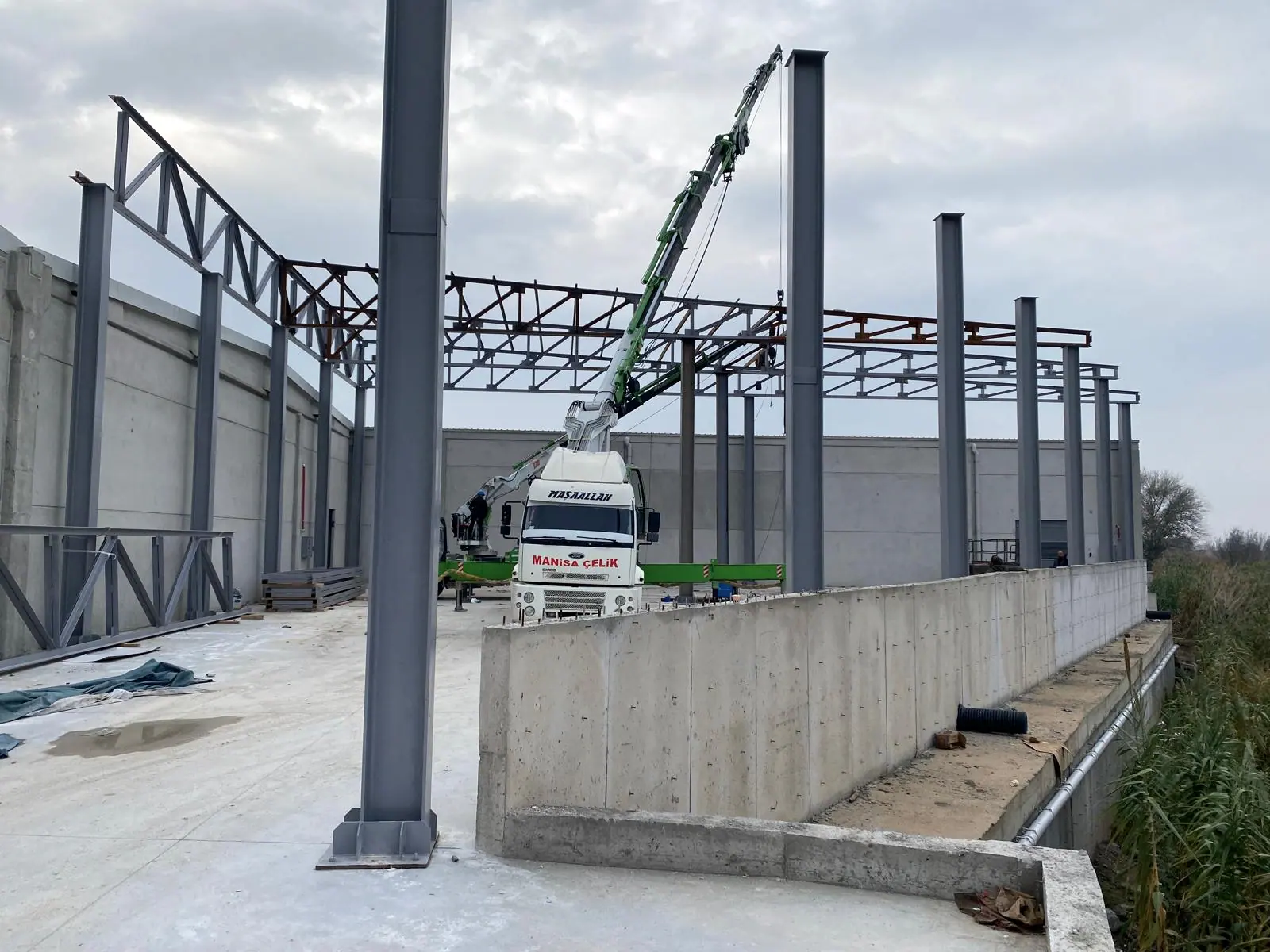 Dikilmiş çelik kolonlar ve ilk kafes makas hattı, kamyon üstü vinç ve önde betonarme istinat duvarı