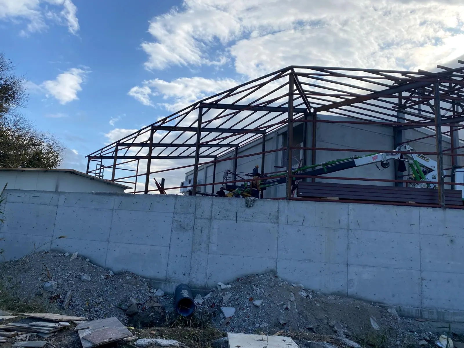 Beton duvarın ardında yükselen çelik çatı makasları, kırmızı sandviç panel istifi ve baretli montaj ekibi