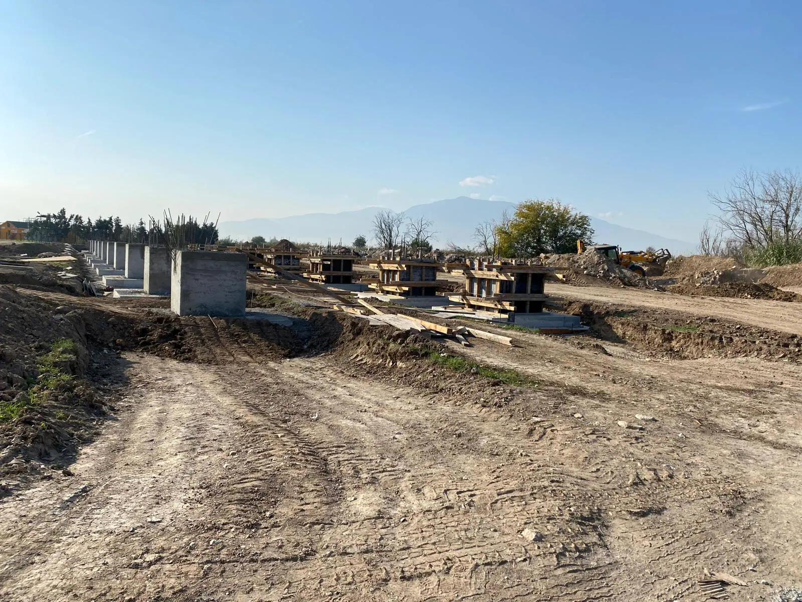 Toprak şantiye yolu boyunca sıralanmış kolon kaideleri, ahşap temel kalıpları ve arkada dağ silsilesi