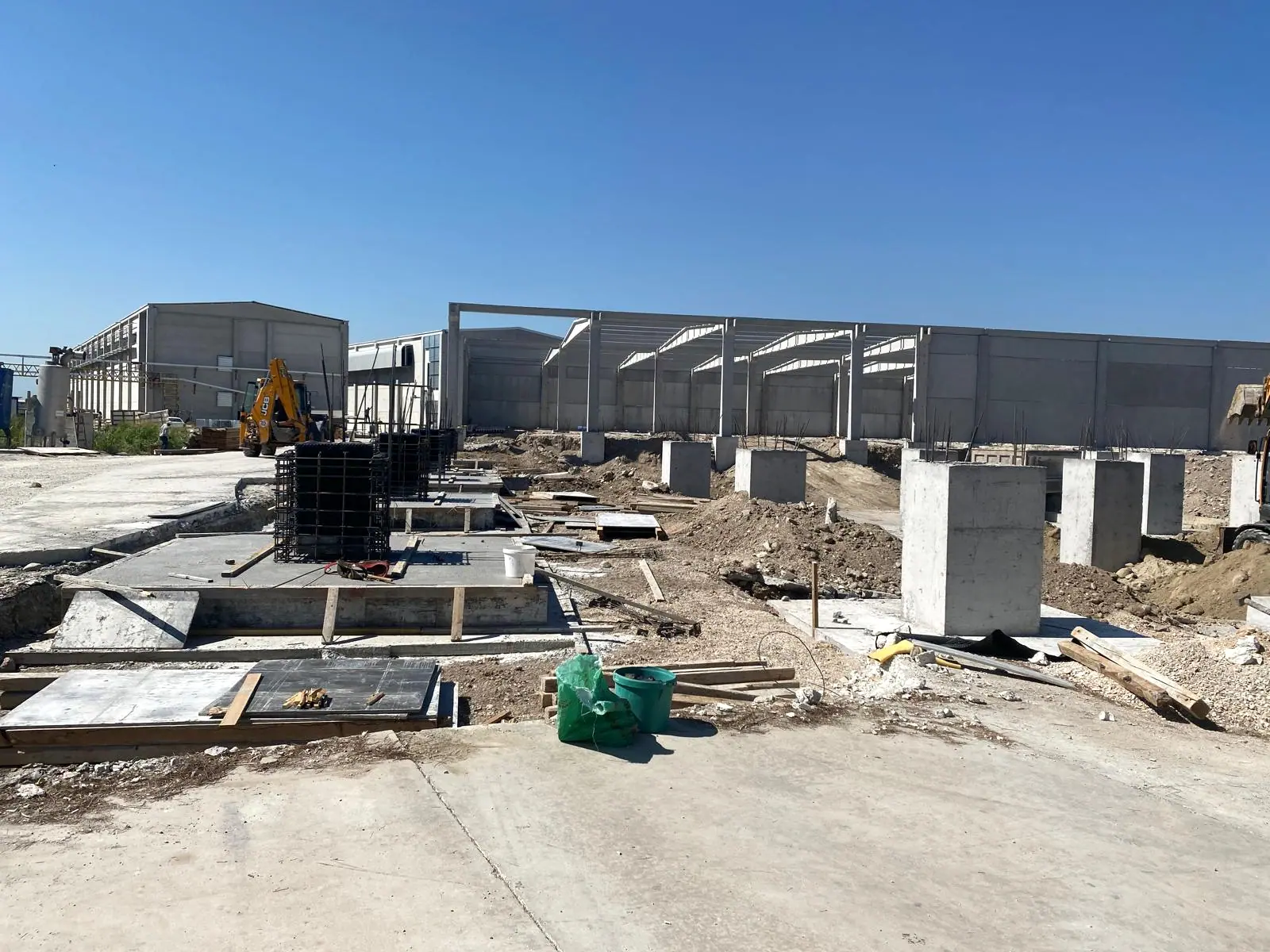 Saha betonu üzerinde temel kalıp panelleri, siyah sargılı kolon donatıları ve arkada uzun beton panel duvarlı hol