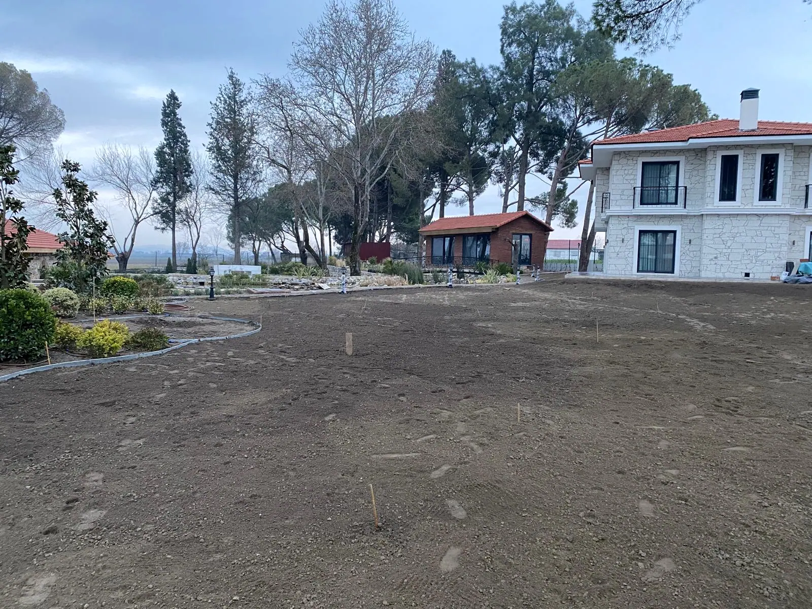 Çim ekimine hazırlanmış tesviyeli toprak zemin, metal bordürlü çalı yatakları ve taş kaplamalı iki katlı yapı