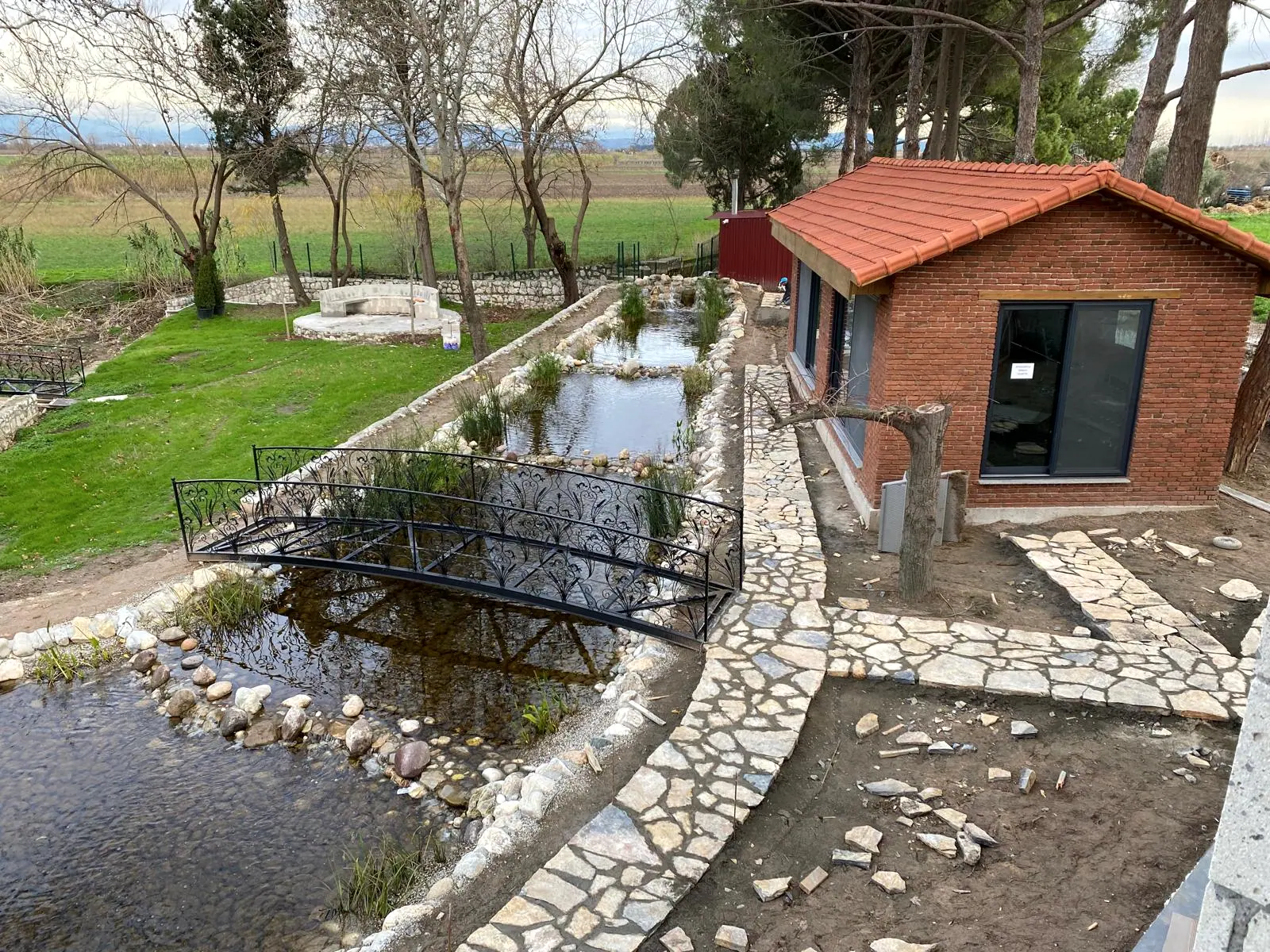 Doğal taş yürüyüş yolu, süs havuzu üzerinde ferforje köprü ve kiremit çatılı tuğla ek yapı