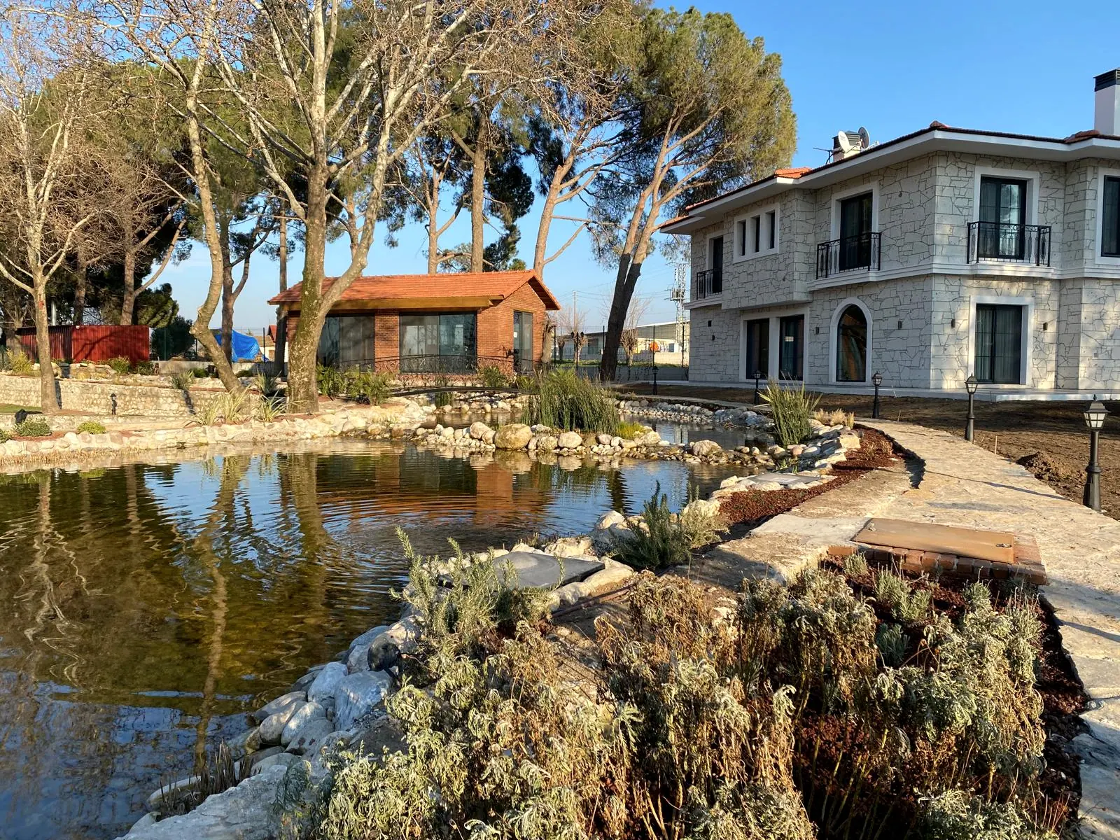 Taş kaplamalı iki katlı villa, önünde süs havuzu, yüksek çam ağaçları ve tuğla bahçe evi