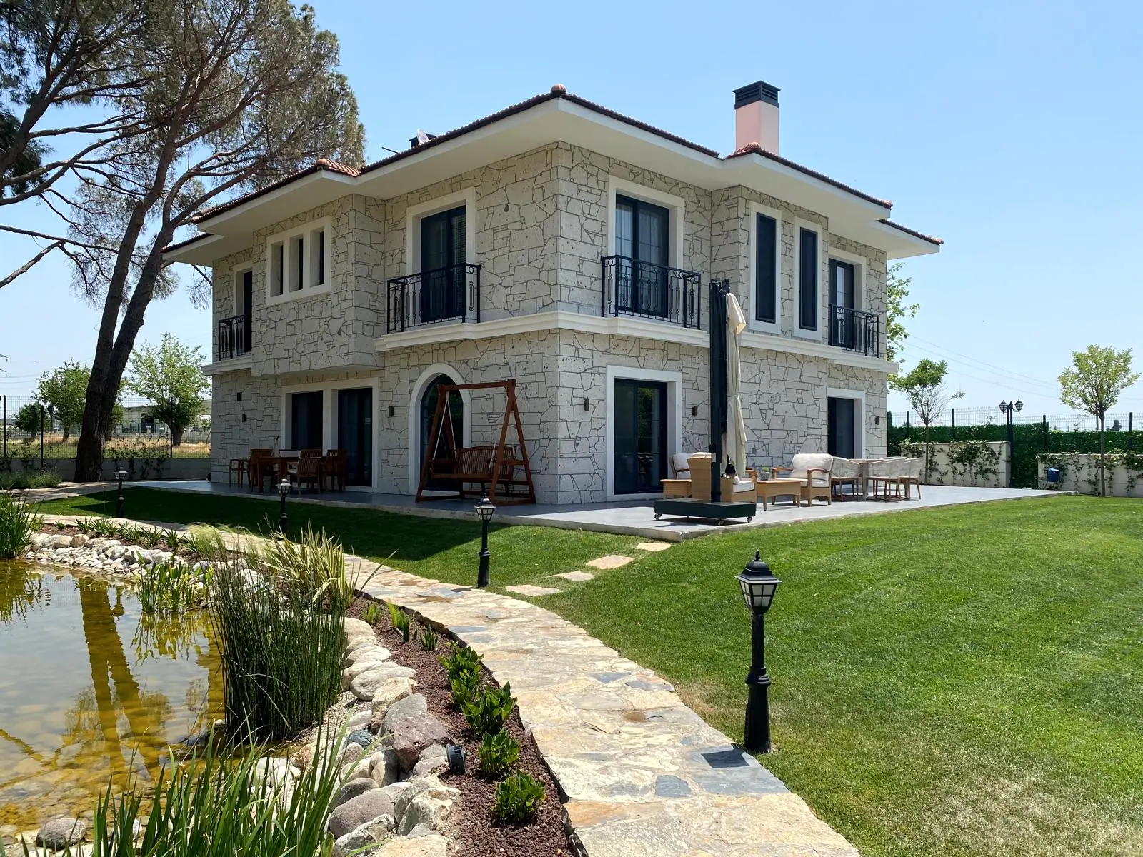 Bakımlı çim, doğal taş yol ve süs havuzu ile çevrelenmiş taş kaplama iki katlı villa